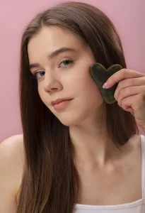 A young woman uses a gua sha tool to massage her face, promoting relaxation and improving circulation.
