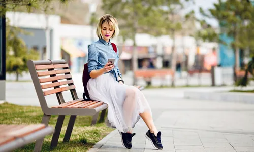 A blonde woman sitting comfortably on a wooden bench while checking her smartphone.