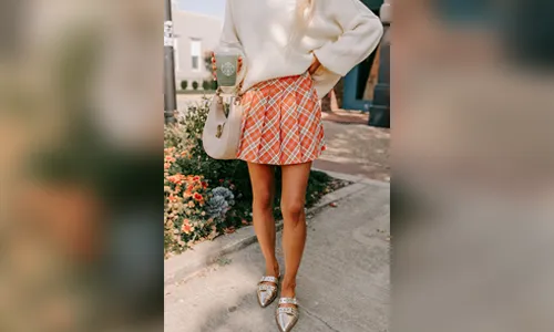 A woman wearing a vibrant orange plaid pleated skort paired with an oversized cream knit sweater and metallic gold pointed-toe buckle flats.